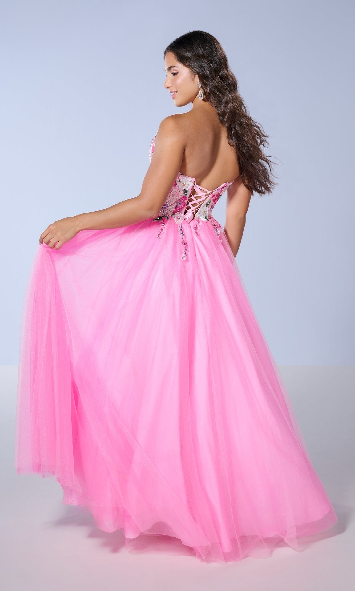 A woman stands turned to the side against a light background, holding out the skirt of the Long Prom Dress: Tiffany 16142, a pink strapless tulle gown with a lace-up back.
